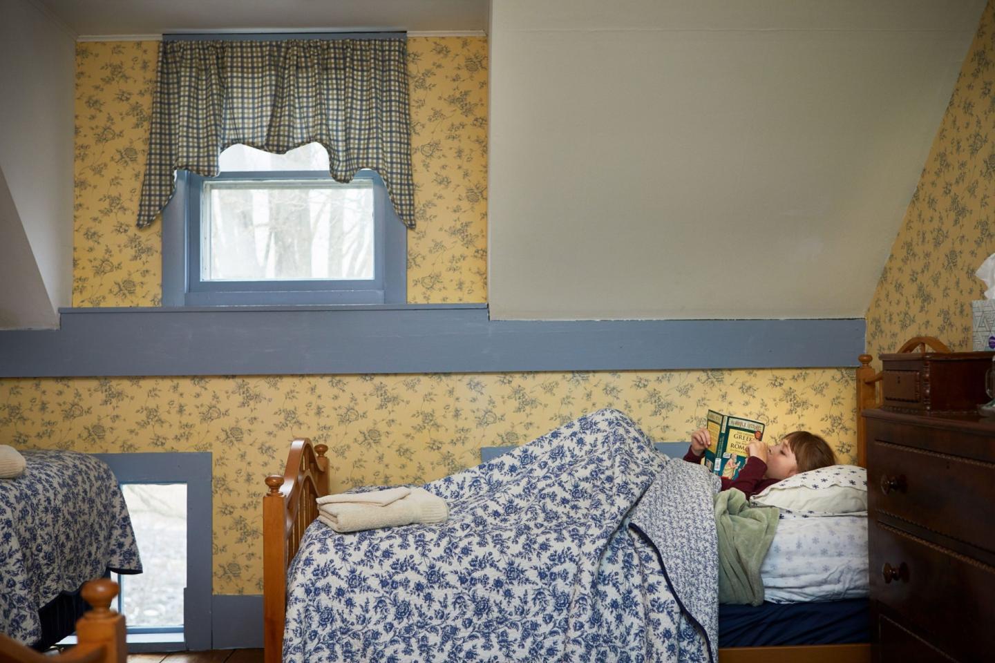 Child reading in a cozy bedroom with floral wallpaper and twin beds.