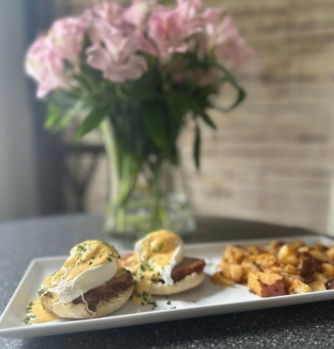 Eggs Benedict and potatoes on a plate, pink flowers in the background.