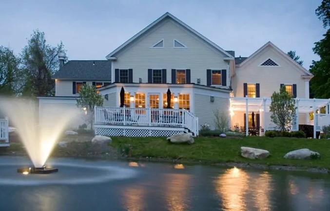 Luxury house by a pond with a lit fountain at dusk.