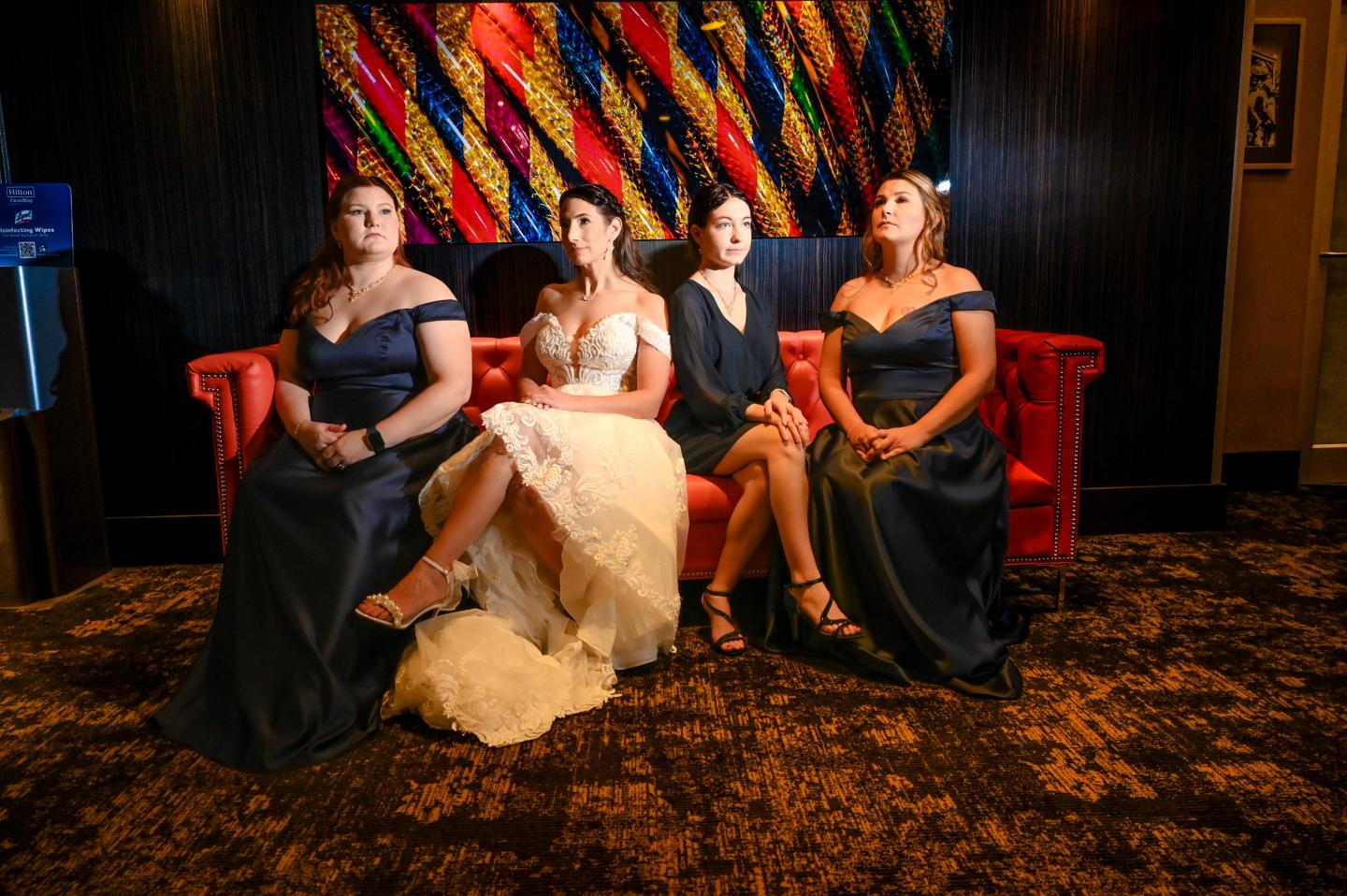 Four women sit on a red couch in formal gowns under colorful wall art.