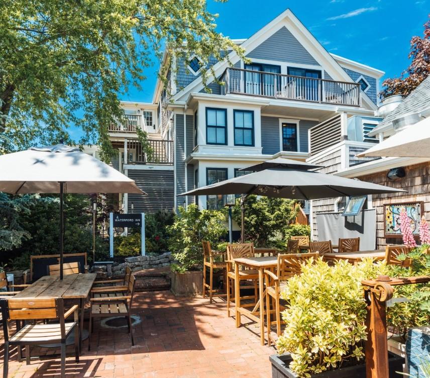 Sunny patio with tables, umbrellas, plants, and a large blue house in the background.