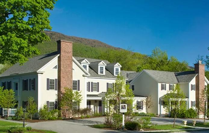 Large white house with green shutters, two chimneys, and surrounding greenery under a blue sky.