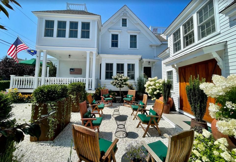 White house with a patio, green chairs, flowers, and American flag, under clear blue sky.