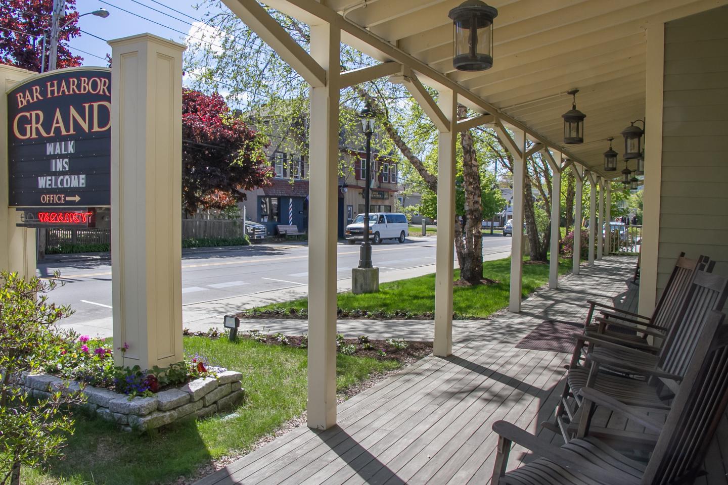 Hotel porch with rocking chairs and hanging lanterns, near a street lined with trees.