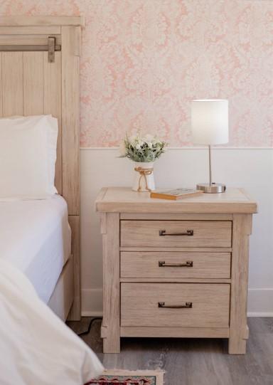 Wooden nightstand with a lamp, flowers, and a book beside a bed.