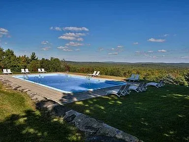 Outdoor pool with lounge chairs, surrounded by greenery under a clear blue sky.