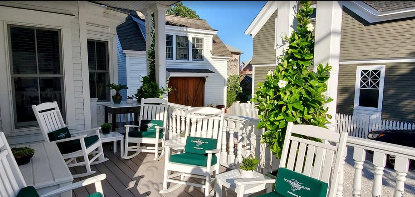 Porch with white rocking chairs, green cushions, and potted plants under a sunny sky.