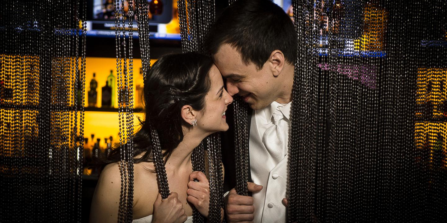 Couple smiling closely behind dark chain curtain, bar setting in background.