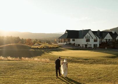 Bride and groom walking across a sunlit field near a large house.