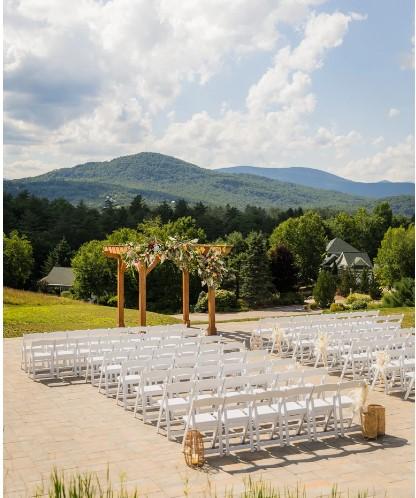 Outdoor wedding setup with chairs and floral arch, mountains in the background.
