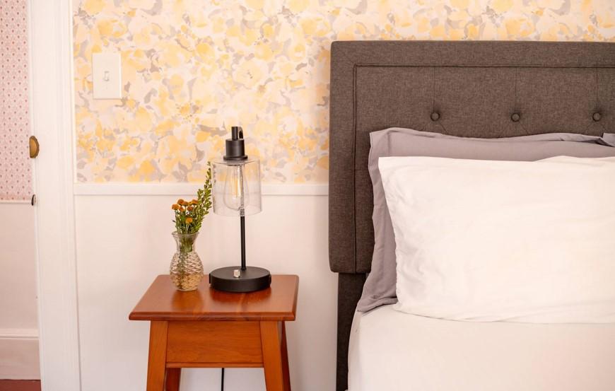 Bedside table with a lamp and flowers, beside a gray headboard against floral wallpaper.