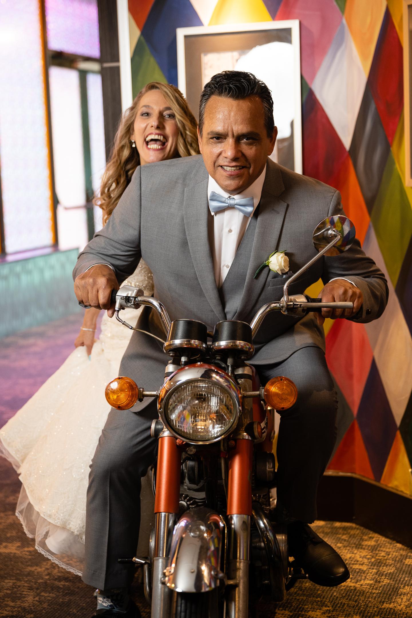 Couple in wedding attire riding a motorcycle indoors, colorful patterned wall behind.