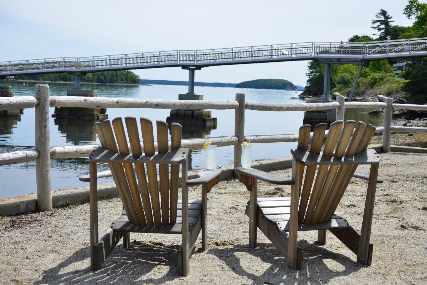 Wooden chairs on a sandy beach facing a pond with a bridge in the background.