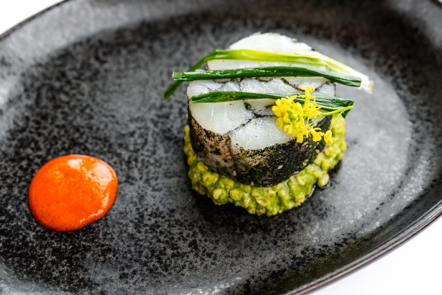 Seared fish on green purée, topped with herbs, next to red sauce on black plate.