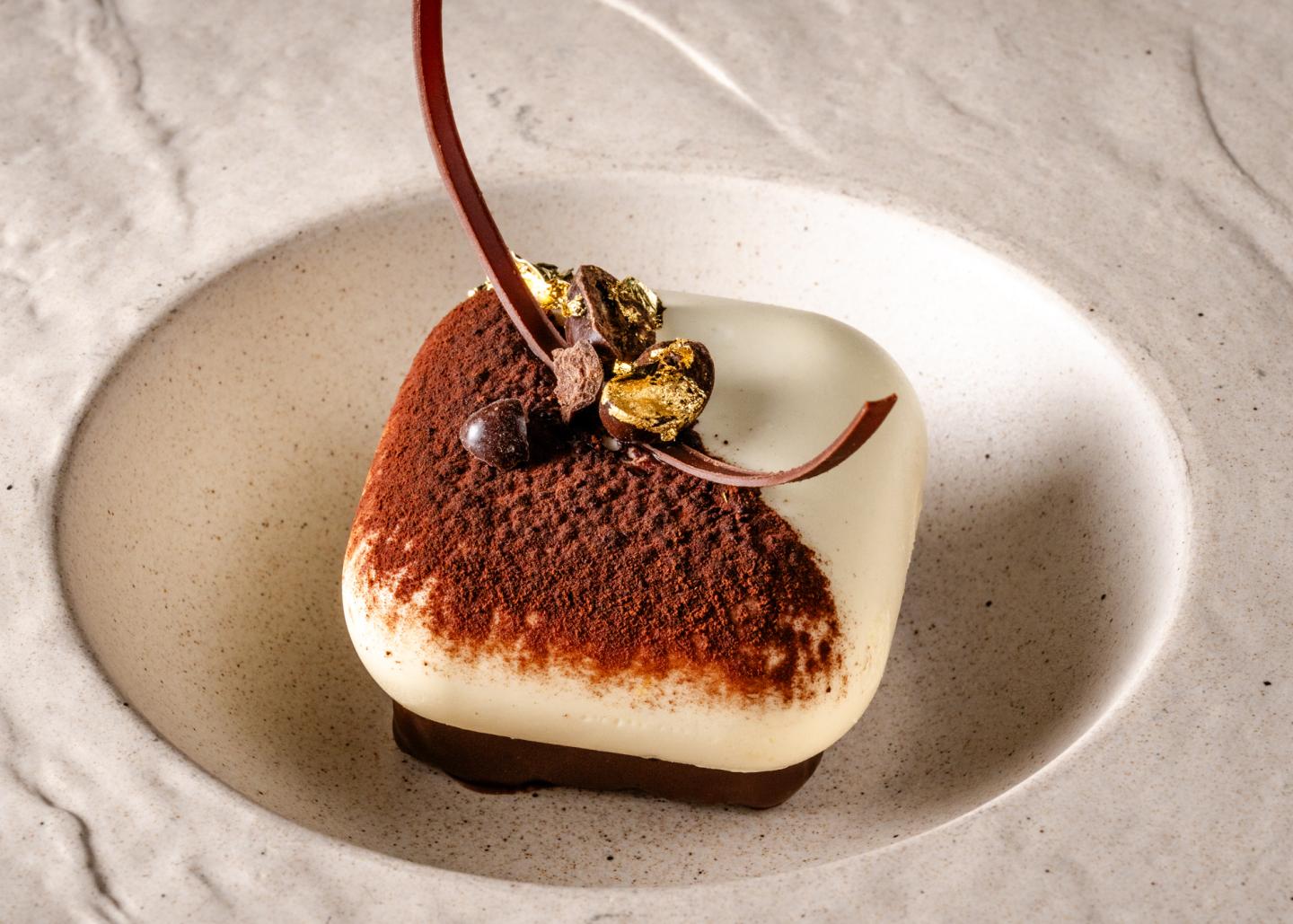 Square dessert with chocolate and cream, garnished with gold leaf on a textured plate.