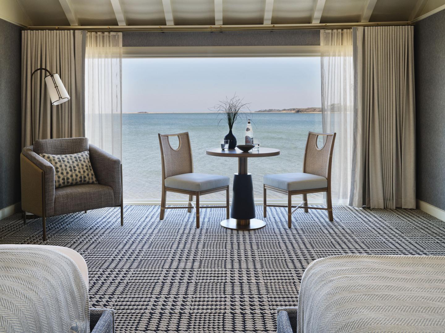 Modern hotel room with ocean view, two chairs, and a round table by a large window.