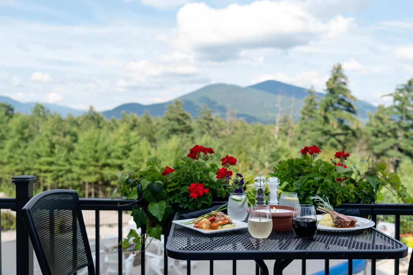 Outdoor dining table with food, drinks, mountain view, and red flowers.