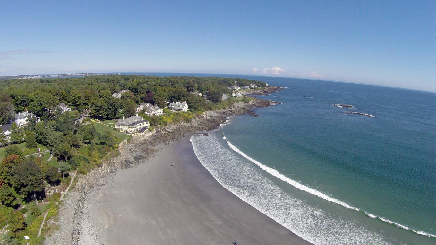 Aerial view of a sandy beach with gentle waves and houses along a green coastline.