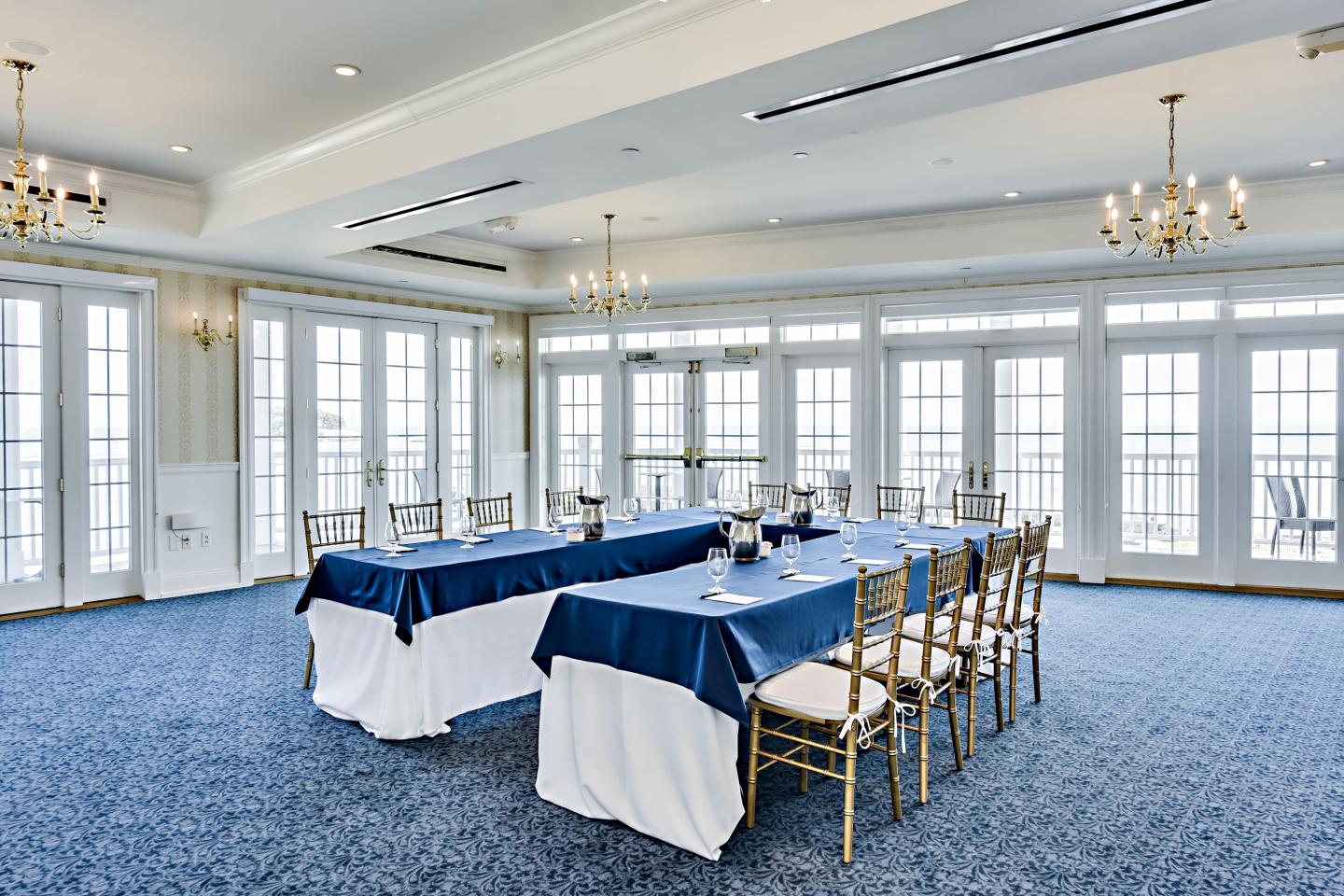Bright banquet room with long tables, blue tablecloths, and many windows.