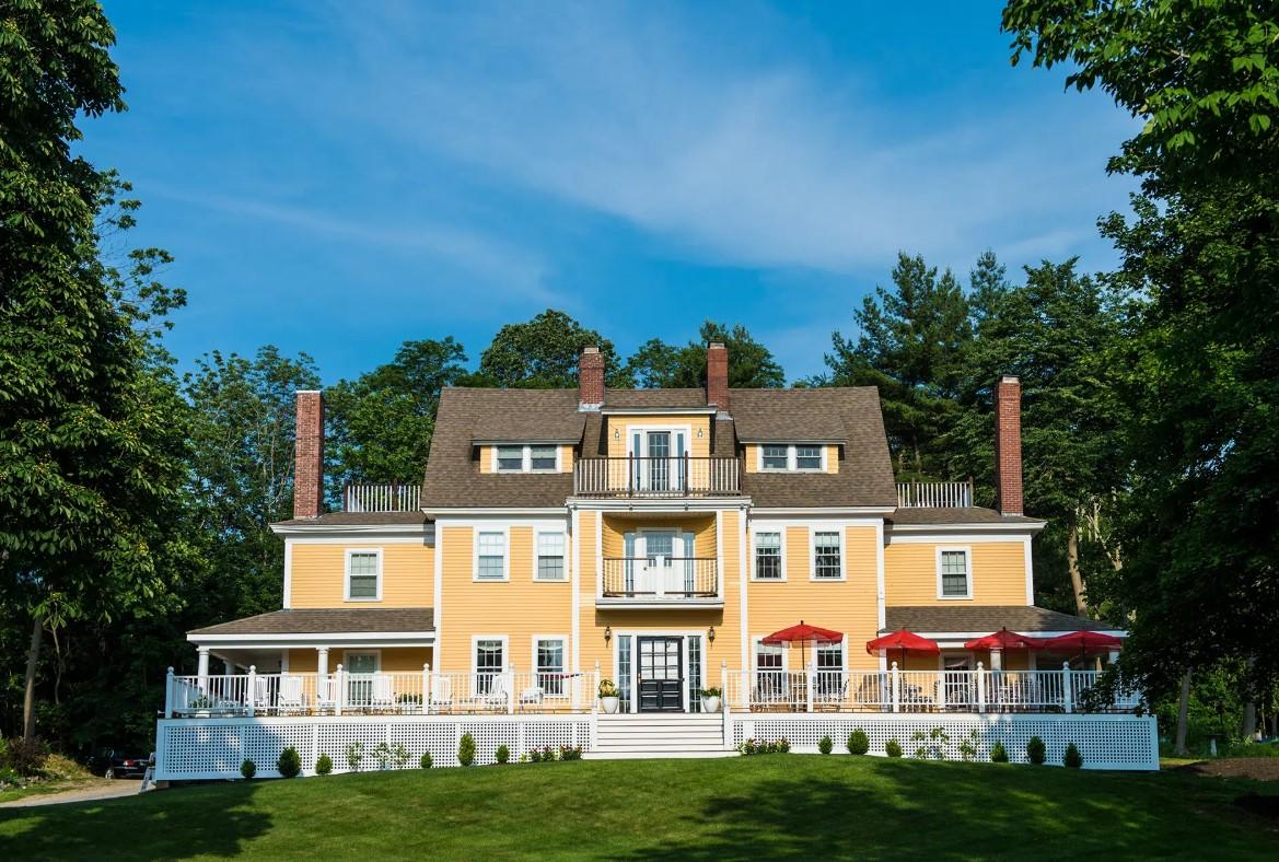 Large yellow house with a white porch, surrounded by trees and a blue sky.