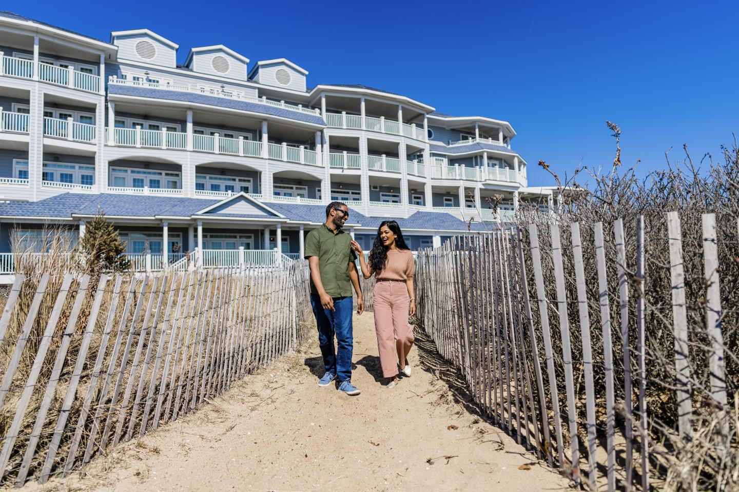 Couple walking on sandy path with beach hotel in background.