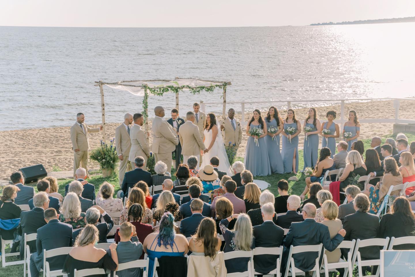 Beach wedding ceremony with bridal party, guests seated under sunny sky.