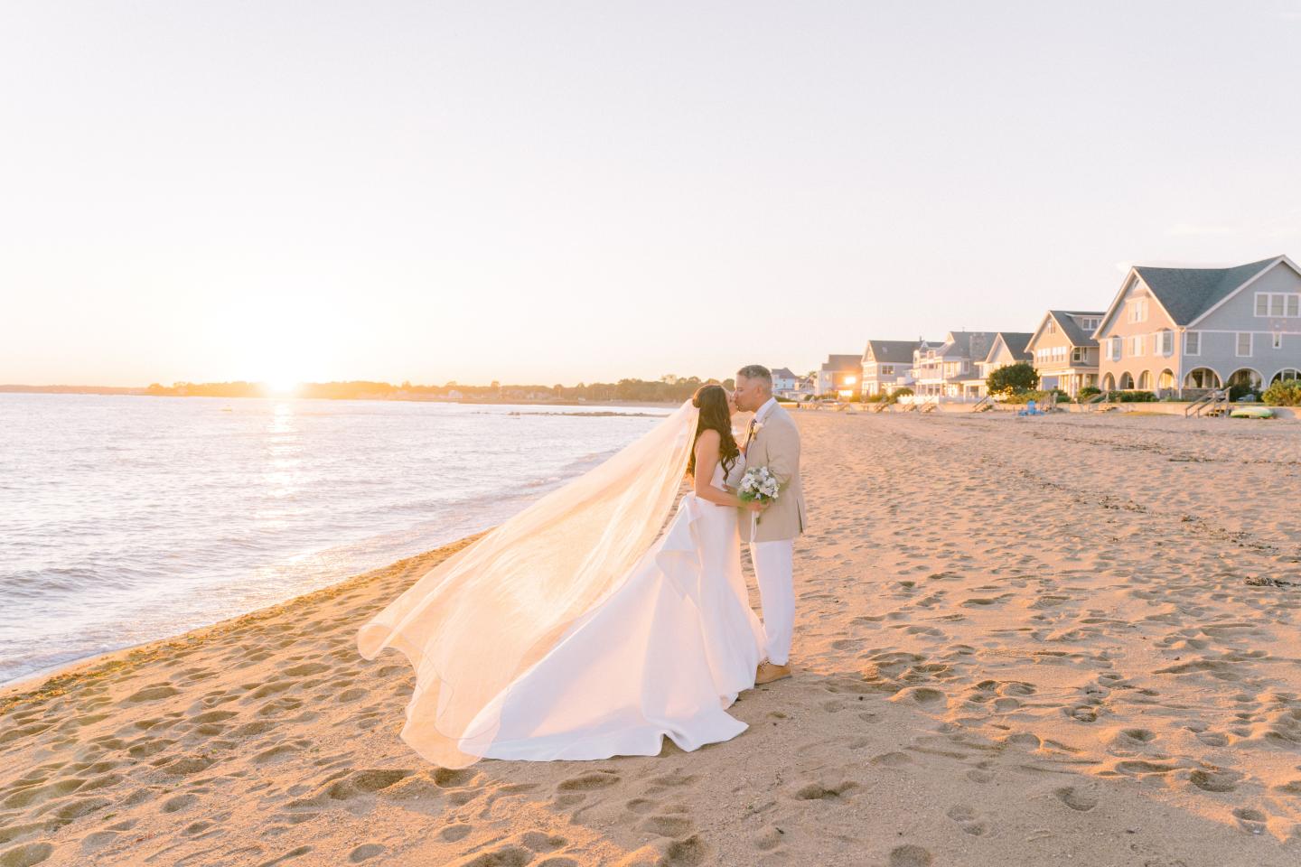 Bride and groom embrace on a sunlit beach at sunset.