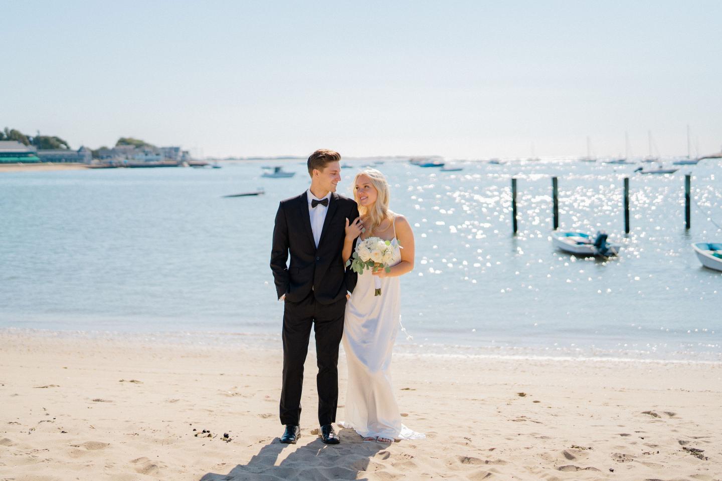 Couple in wedding attire on a sunny beach with boats in the background.