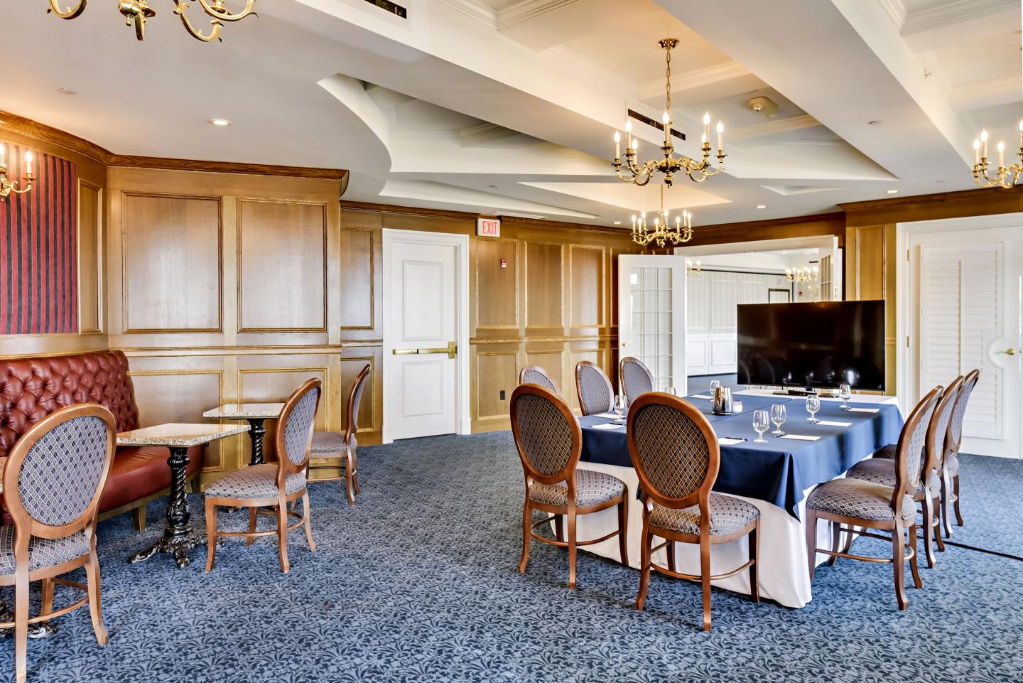 Elegant meeting room with long table, chairs, and large TV. Warm lighting, beige walls, and blue carpet.