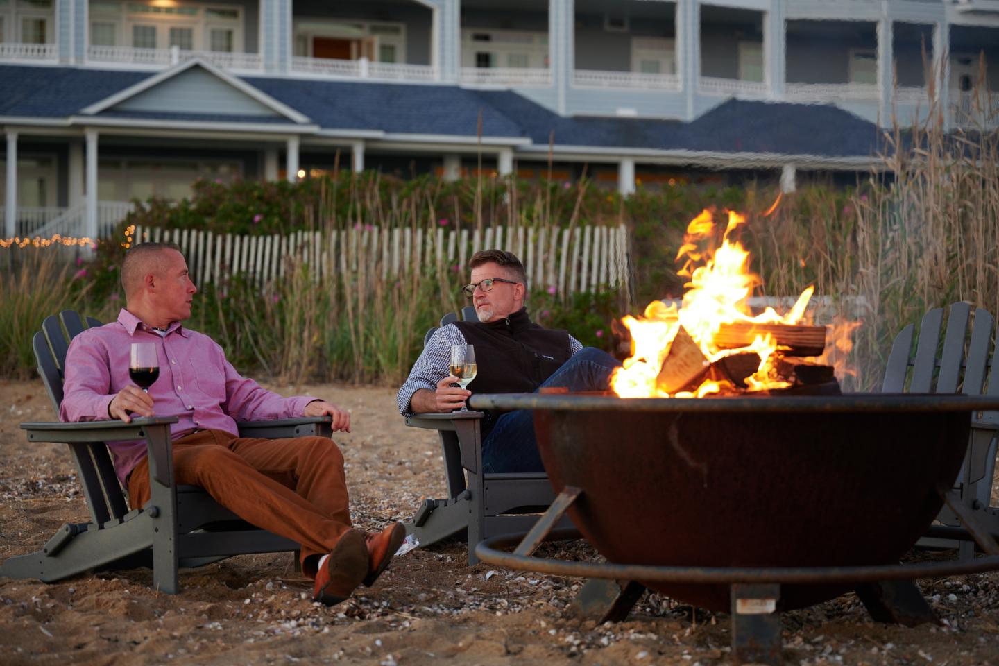Two people sitting by a beach fire pit, holding wine glasses, near a house.