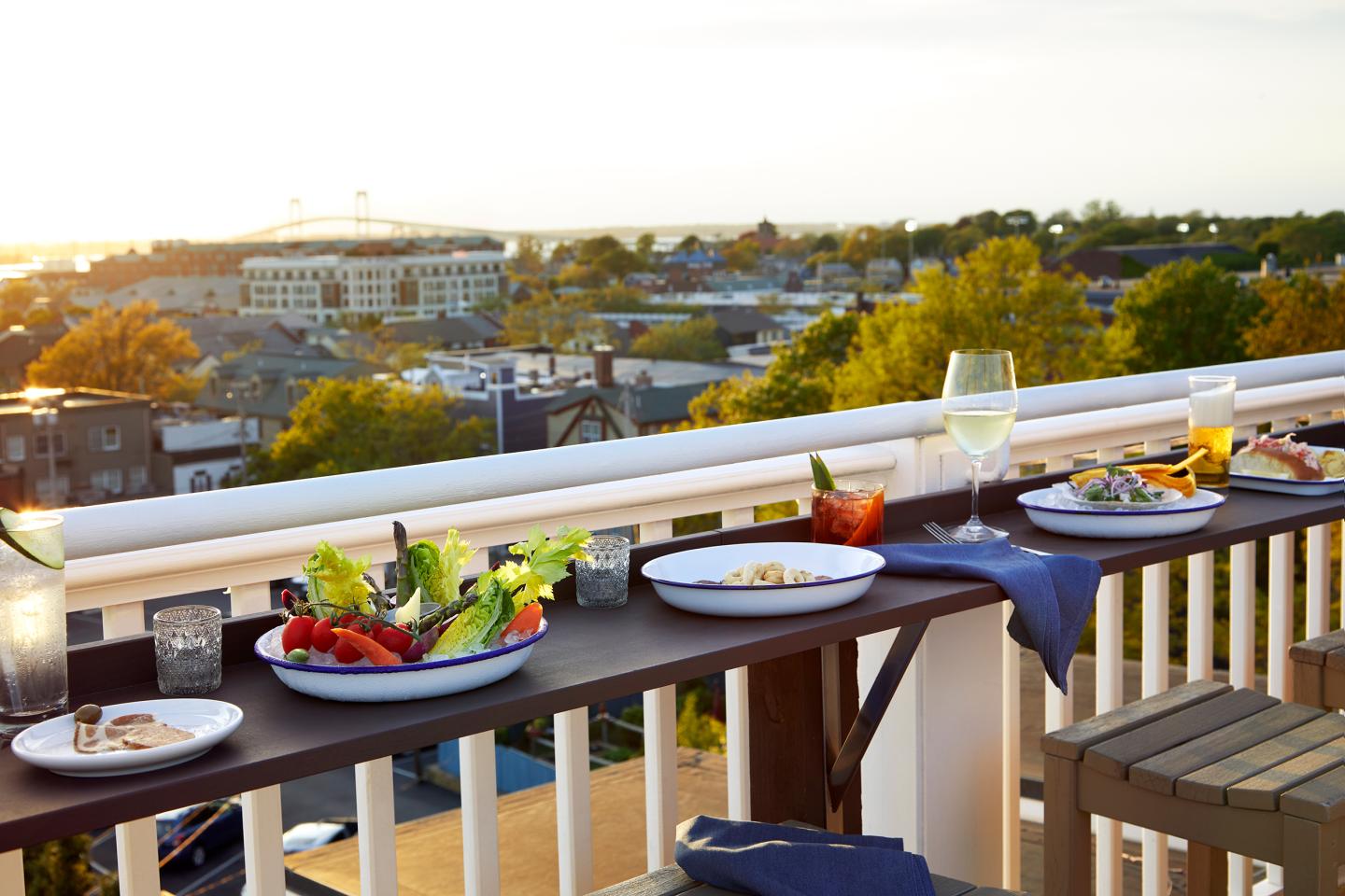 Rooftop table with drinks and plates of food, overlooking a cityscape at sunset.