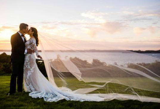 Bride and groom kissing at sunset by a lake, flowing veil, elegant attire.