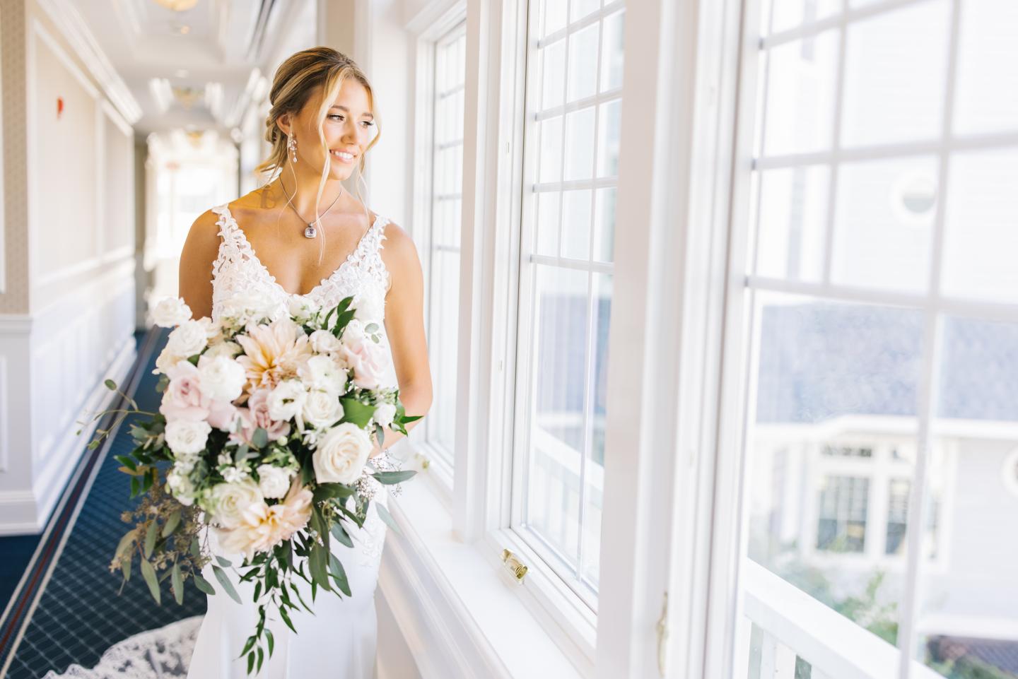 Bride in white dress holding bouquet, standing by large windows, smiling.