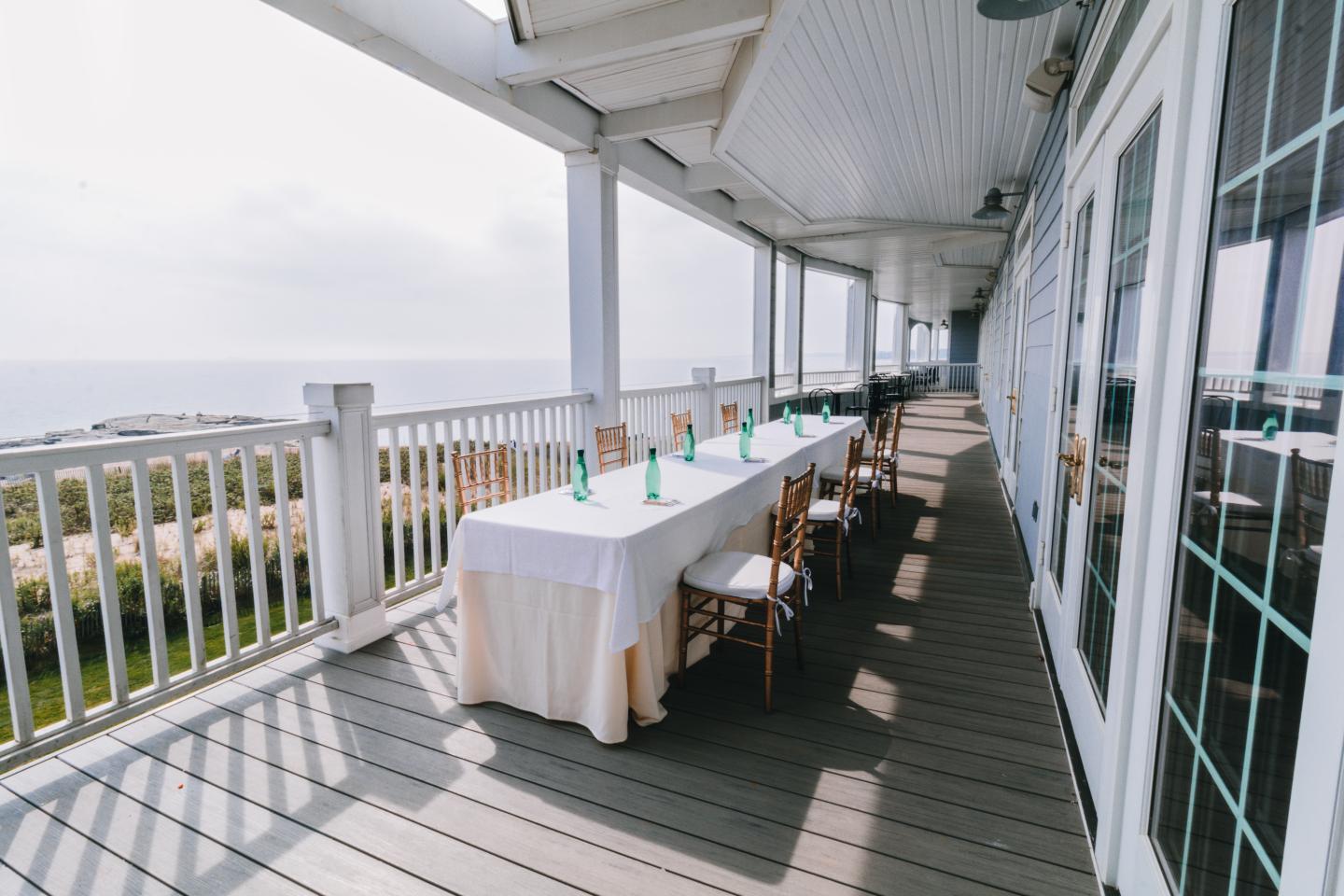 Long table on a sunny porch, set with bottles, overlooking greenery and sea.