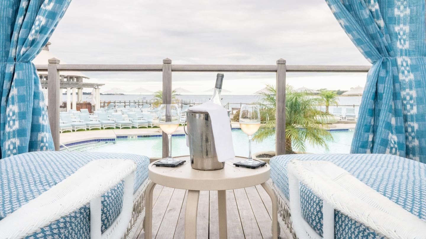 Poolside cabana with blue curtains, bottles, and glasses; overlooking a pool and palm trees.