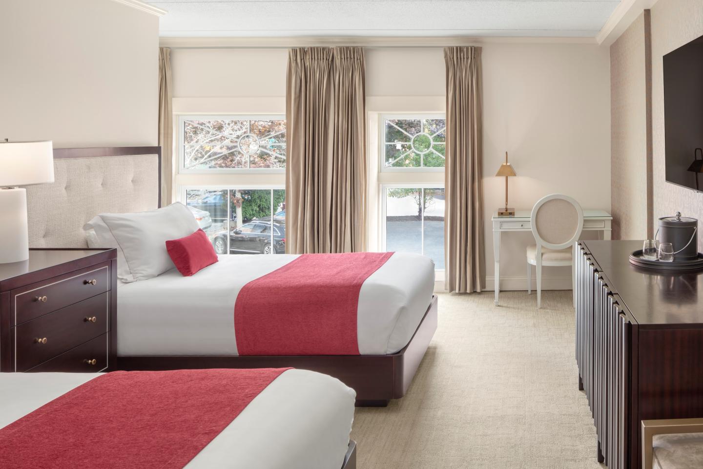 Elegant hotel room with two double beds, red accents, and a desk by the window.