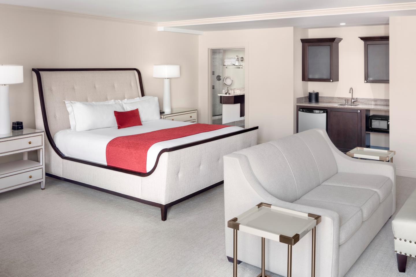 Spacious hotel room with bed, sofa, and kitchenette. Neutral tones with a red accent pillow.