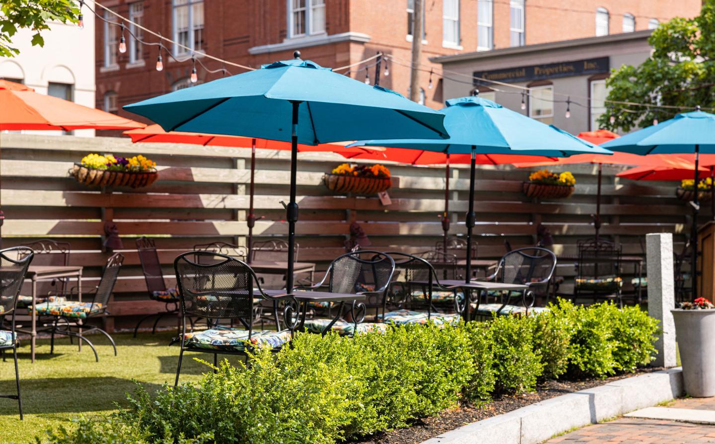 Outdoor patio with blue umbrellas, metal chairs, and potted plants, on a sunny day.