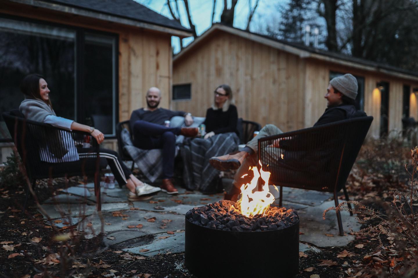 Group of people sitting around an outdoor fire pit, surrounded by autumn leaves.