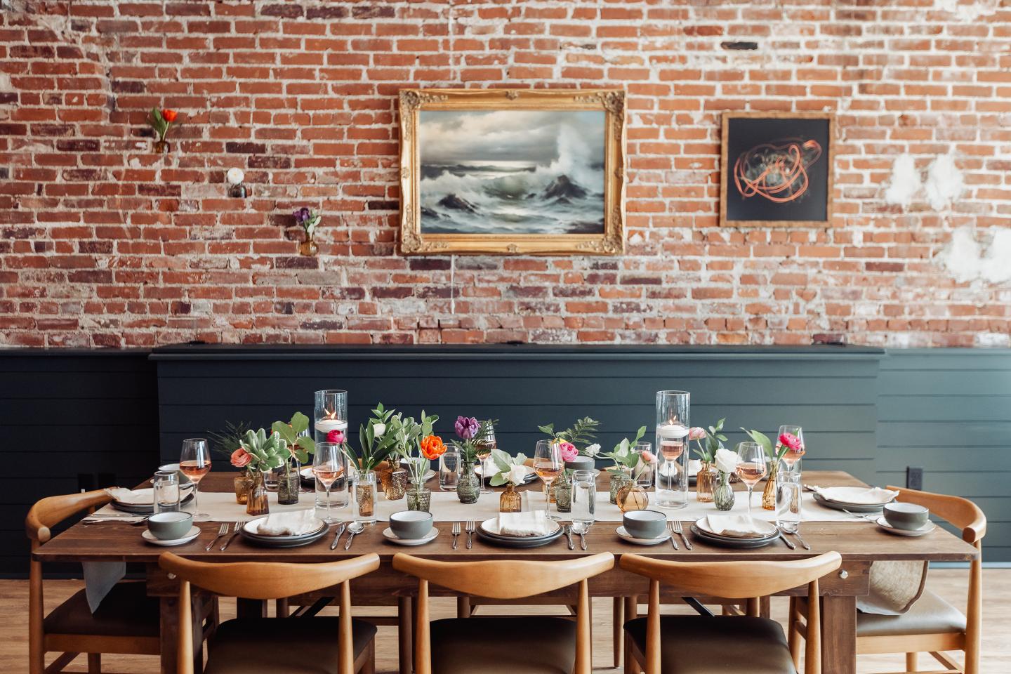 Dining table set with flowers, brick wall background, hanging artworks.