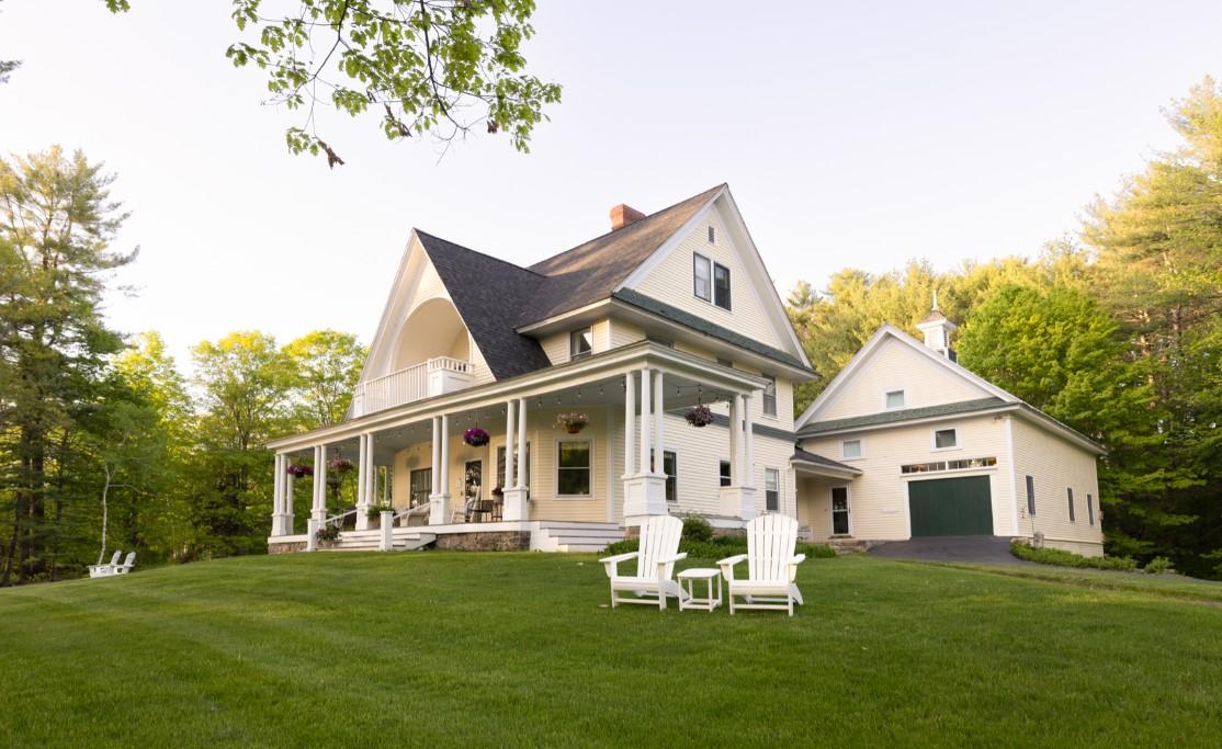 White house with porch and chairs on a grassy lawn, surrounded by trees.