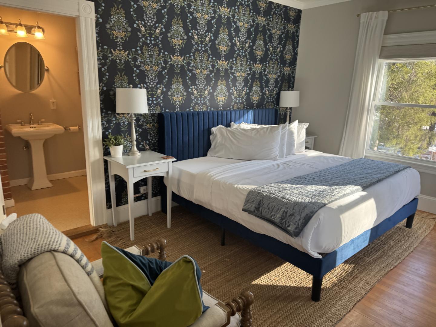Cozy bedroom with a blue bed, floral wallpaper, and a view into a bathroom.