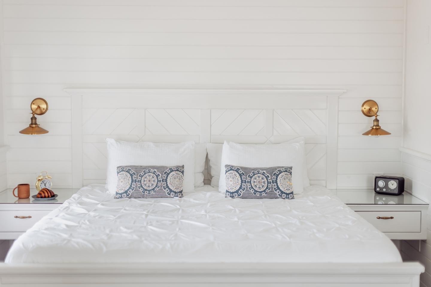 White bed with patterned pillows, flanked by vintage lamps and a clock on side tables.