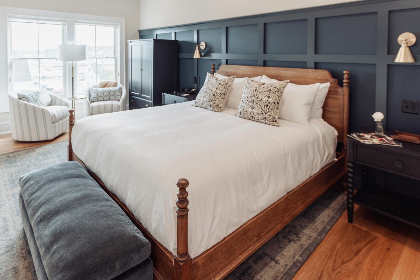 Wooden bed with white linens, navy accent wall, and cozy armchairs by the window.