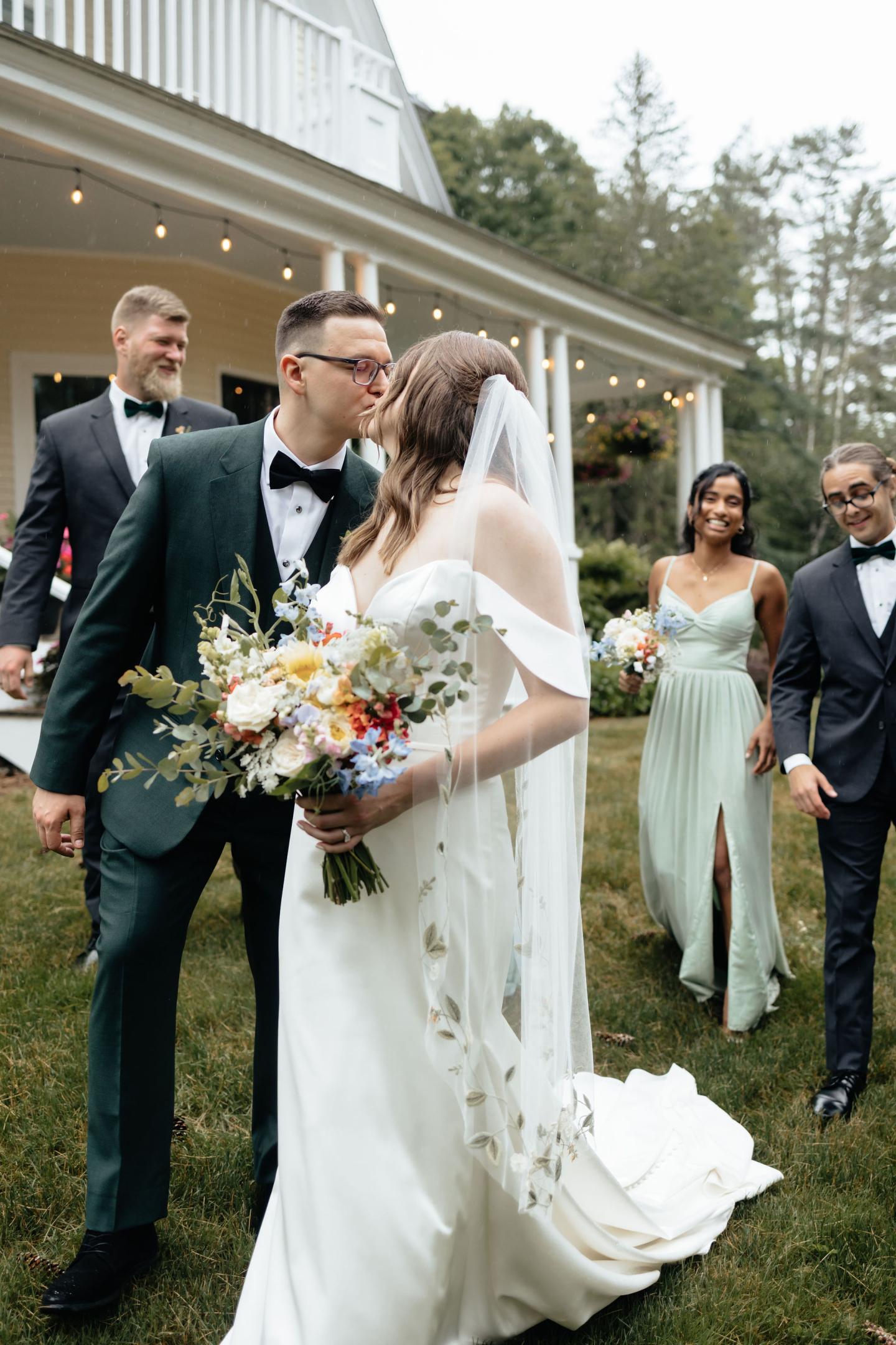 Bride and groom kissing, surrounded by smiling wedding party outdoors.