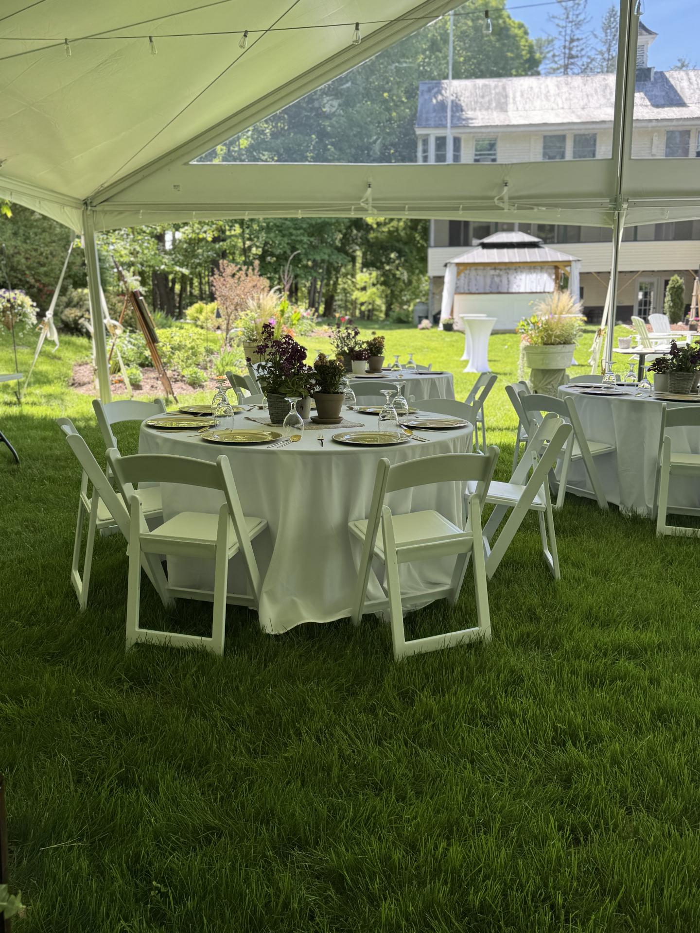 Outdoor event with round tables, white chairs, and floral centerpieces under a canopy.