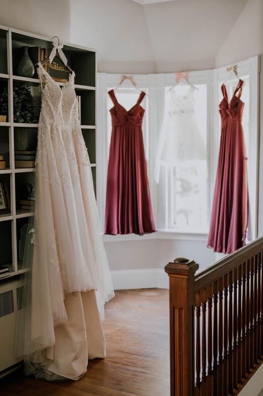 White wedding dress and burgundy bridesmaid dresses hanging by a window.