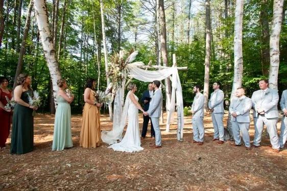 Bride and groom hold hands under a canopy in a forest, surrounded by their wedding party.