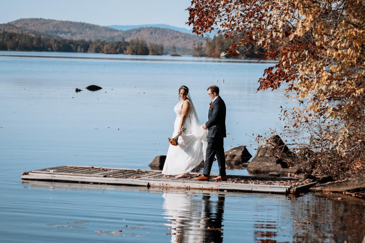 Bride and groom on dock by lake, surrounded by autumn trees.