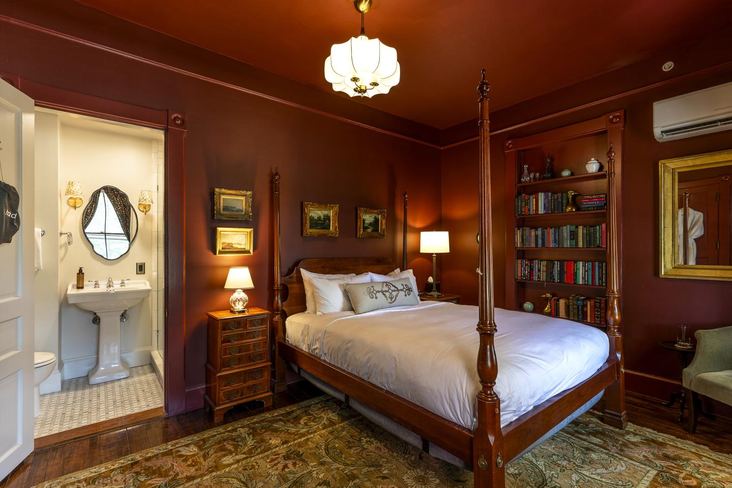 Cozy bedroom with dark red walls, four-poster bed, bookshelves, and a small adjacent bathroom.